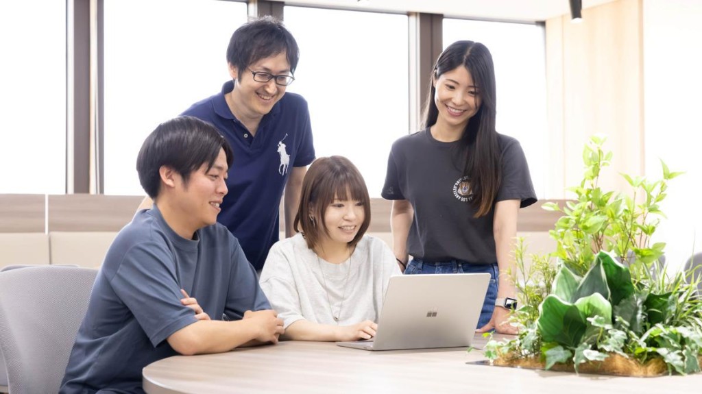 Nhóm nhân viên trao đổi quanh laptop trong văn phòng sáng, có cây xanh. Ảnh minh họa, không phải đội ngũ K.System.